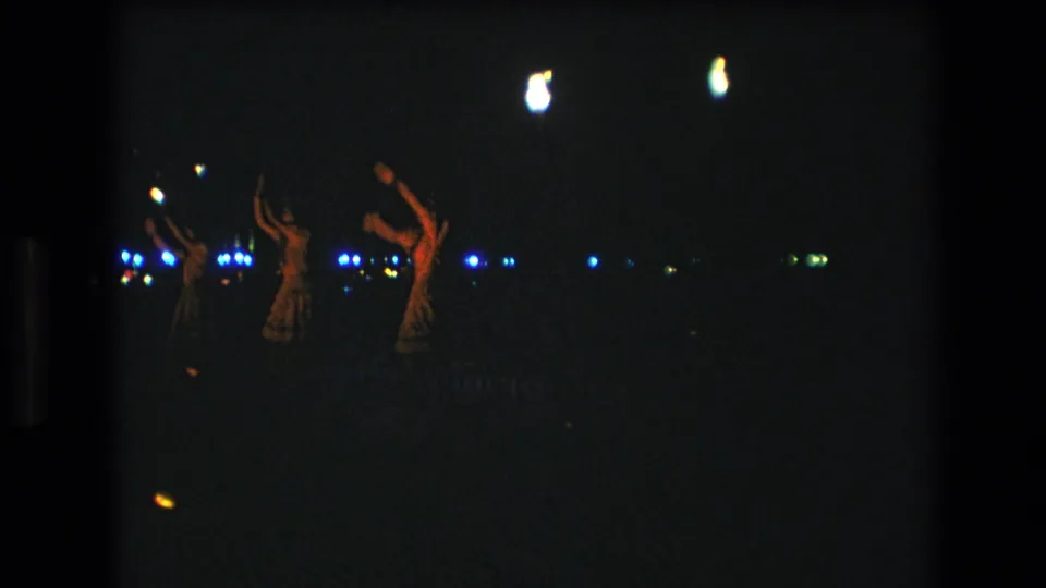 1977 Honolulu frame with several distant performers raising their arms during a nighttime luau-style dance, seen through grainy 8mm film exposure.