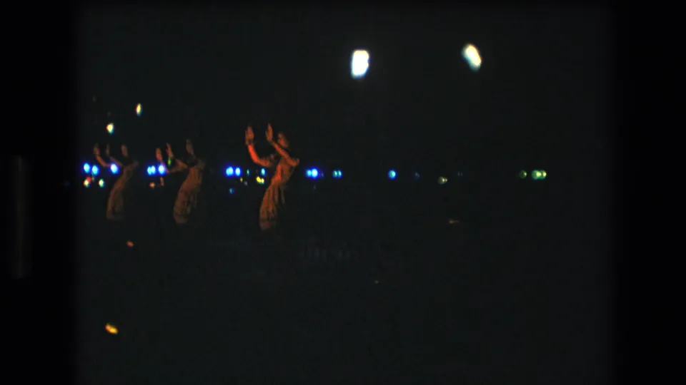 Grainy 1977 Honolulu 8mm frame showing distant dancers continuing a nighttime luau-style performance amid scattered blue lights and warm firelit highlights.