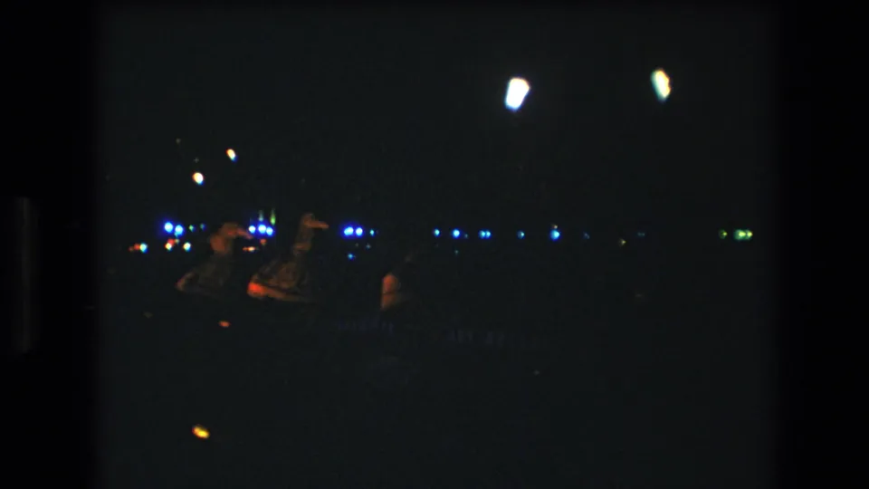Final 1977 Honolulu frame showing a distant dancer bending low during a nighttime luau-style scene, with torchlike highlights and scattered lights across the background.