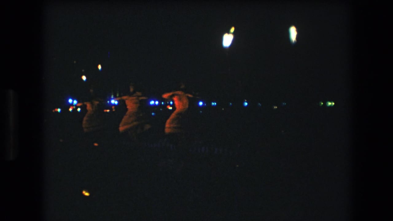 Three women appear to perform hula at night in Honolulu, Hawaii, in 1977 home-movie footage, seen from an audience distance with scattered blue and white lights in the background.