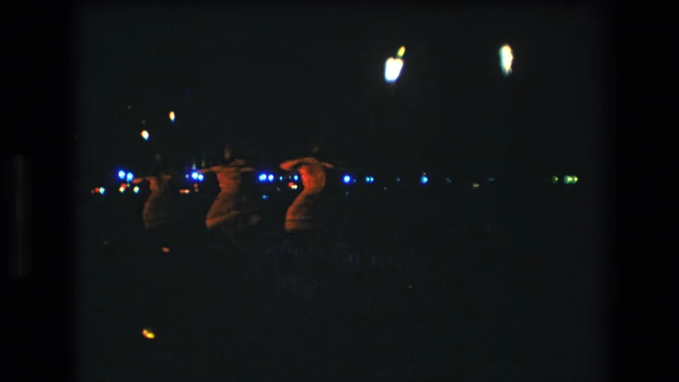 Three women appear to perform hula at night in Honolulu, Hawaii, in 1977 home-movie footage, seen from an audience distance with scattered blue and white lights in the background.