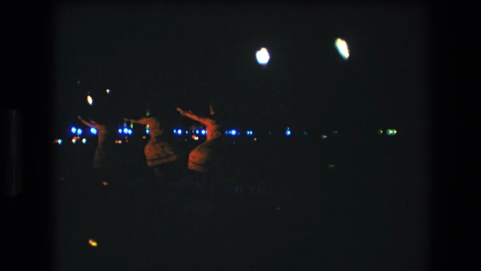 A 1977 Honolulu 8mm frame shows three women dancing hula in a dark outdoor setting, with soft silhouettes, warm clothing tones, and blue lights across the background.