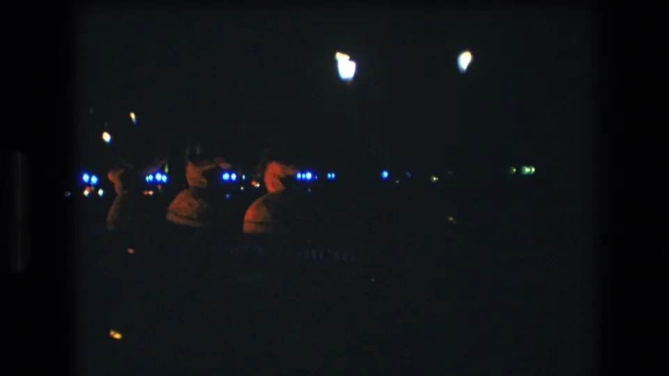 Three women continue a hula dance in 1977 Honolulu home-movie footage, filmed in very low light with grain, soft focus, and scattered blue and white background lights.