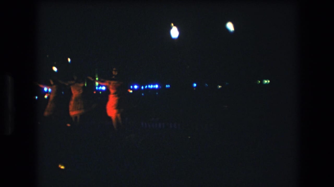 1977 Honolulu-area 8mm frame showing three women dancing at night, with red-orange light on one figure and small blue lights in the background.