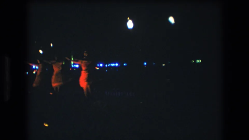 1977 Honolulu-area 8mm frame showing three women dancing at night, with red-orange light on one figure and small blue lights in the background.