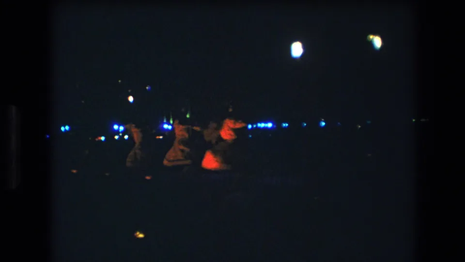 1977 Honolulu 8mm footage frame showing three women dancing at a nighttime event, partly silhouetted against distant blue lights.