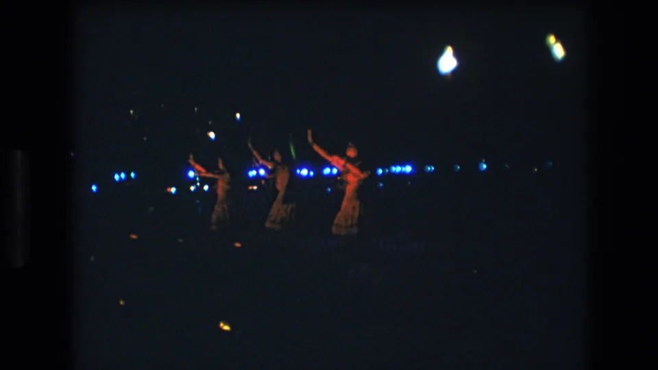 Wide dark 8mm frame from 1977 Honolulu showing women dancing beneath scattered bright lights, with strong blue points across the background.