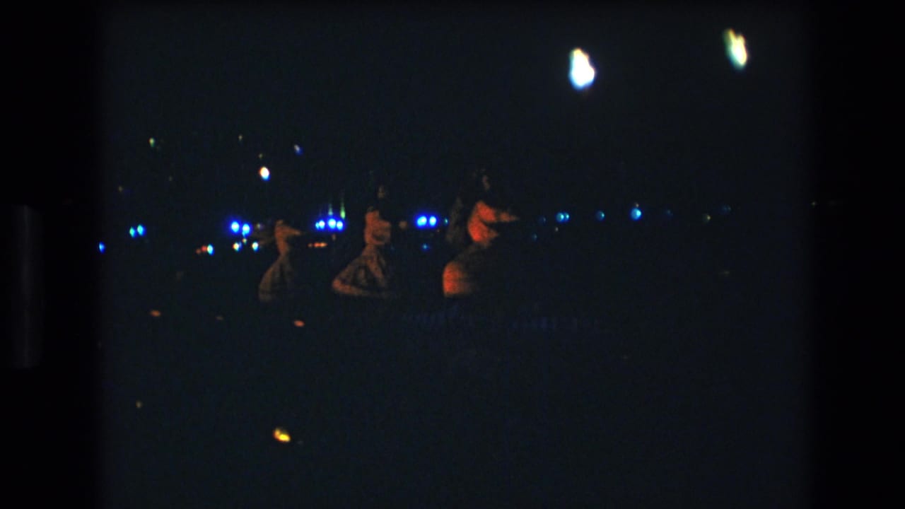 Three distant women dance at night under scattered lights on grainy 8mm film.