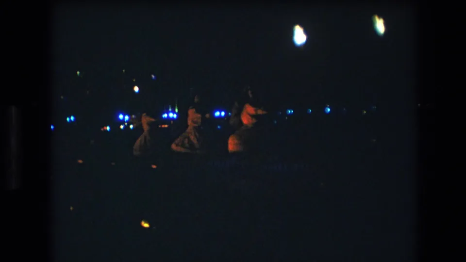 In 1977 Honolulu, Oahu, three women appear to perform a nighttime dance while distant blue-white lights and overhead flares glow in the dark background.