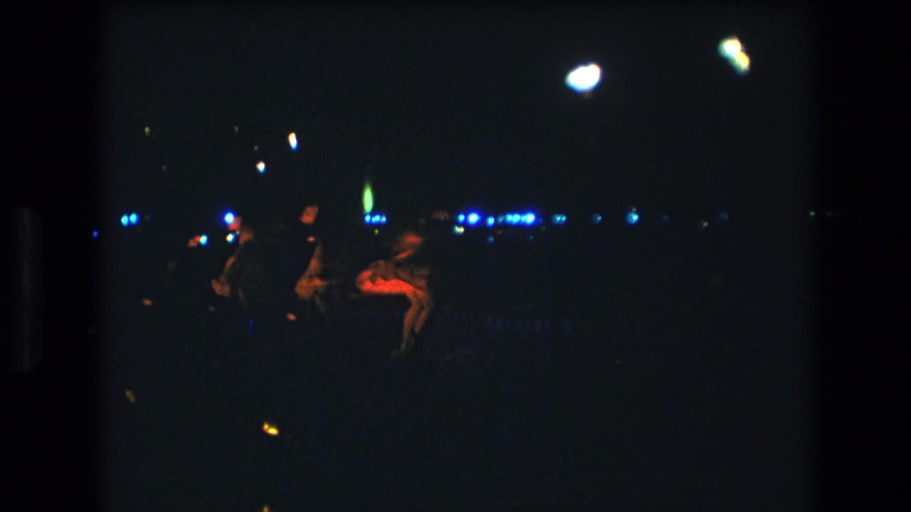 Dancers move across a dark nighttime scene with bright colored fabric and distant lights.