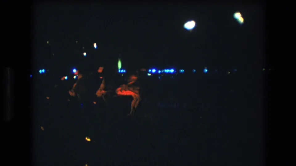 A low-light 8mm frame from Honolulu, Oahu shows women dancing at night, with red fabric catching the light against a dark field of blue and white points.