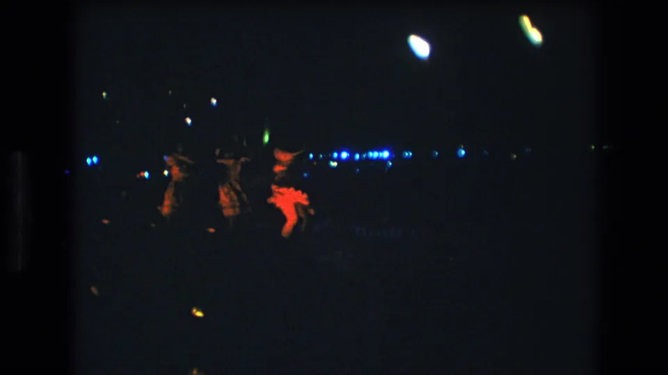 The frame shows three women appearing to gesture in unison during a nighttime dance presentation in 1977 Honolulu, with red fabric and distant lights visible through heavy film grain.