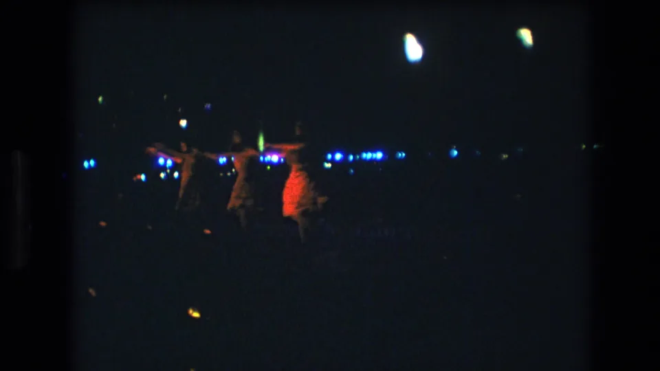 Three women continue the nighttime dance in Honolulu, Oahu, with one red garment especially bright against the dark background and a line of small blue-white lights behind them.