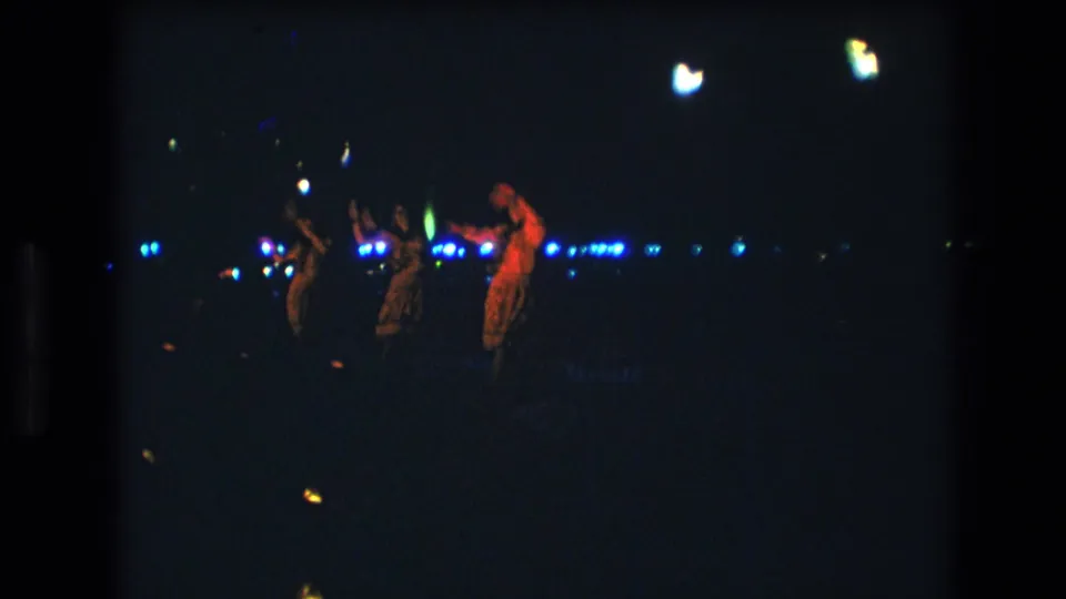 Three women are visible in a nighttime Honolulu performance frame, with one red-lit dancer on the right and two dimmer figures near the center.