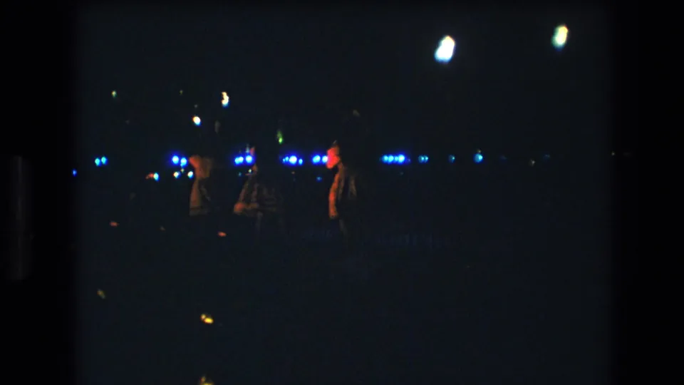 The frame shows the dancers farther into motion on a dark Honolulu stage, with blue points of light behind them and red illumination catching one figure.