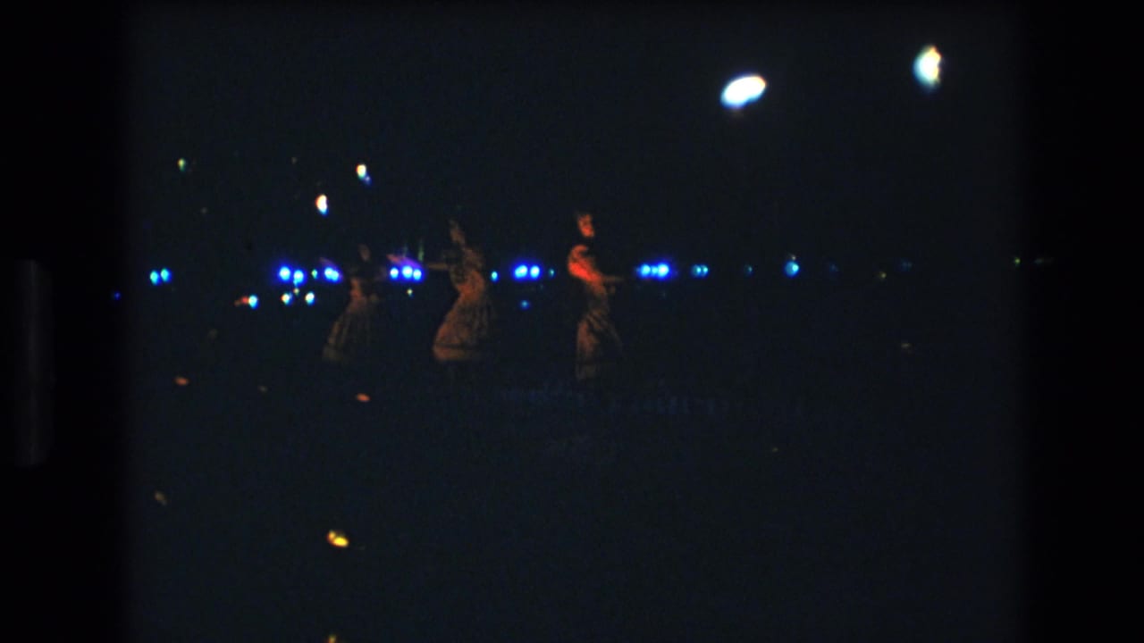 1977 Honolulu home-movie frame showing female dancers at a distance on a dark stage with blue lights and glowing handheld props.