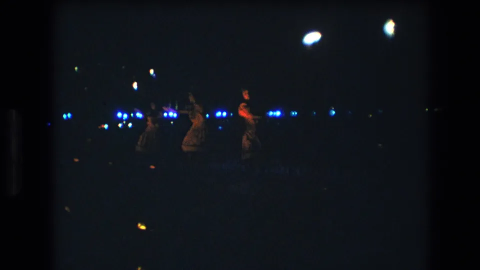 1977 Honolulu home-movie frame showing female dancers at a distance on a dark stage with blue lights and glowing handheld props.