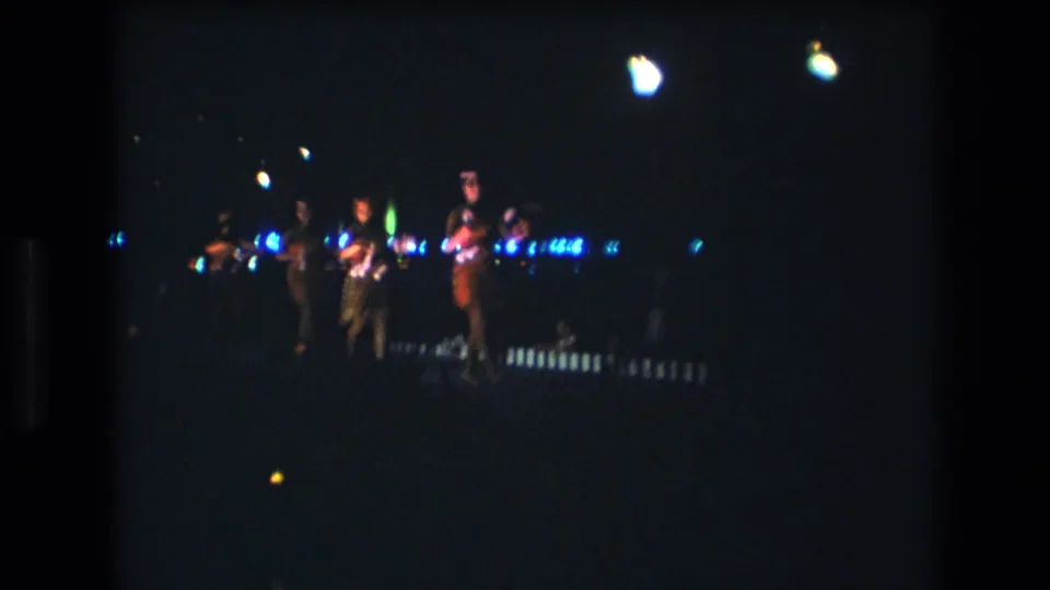 1977 Honolulu 8mm frame showing performers on a dim stage, with bright overhead light bloom and small blue lights across the background.