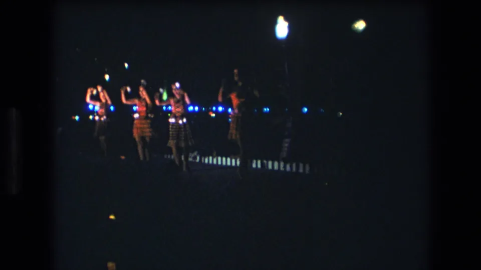 1977 Honolulu home-movie frame showing female dancers in patterned skirts moving with raised arms under blue stage lighting.