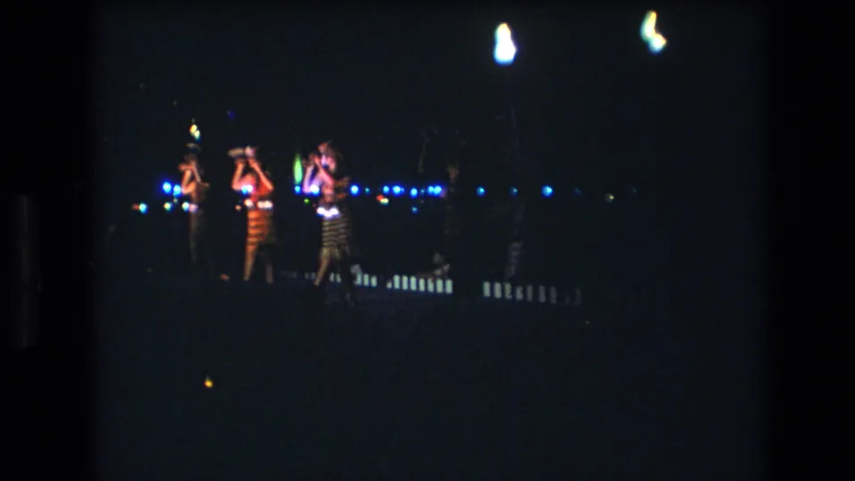 1977 Honolulu 8mm frame showing dancers holding small glowing lights while performing on a dim nighttime stage.