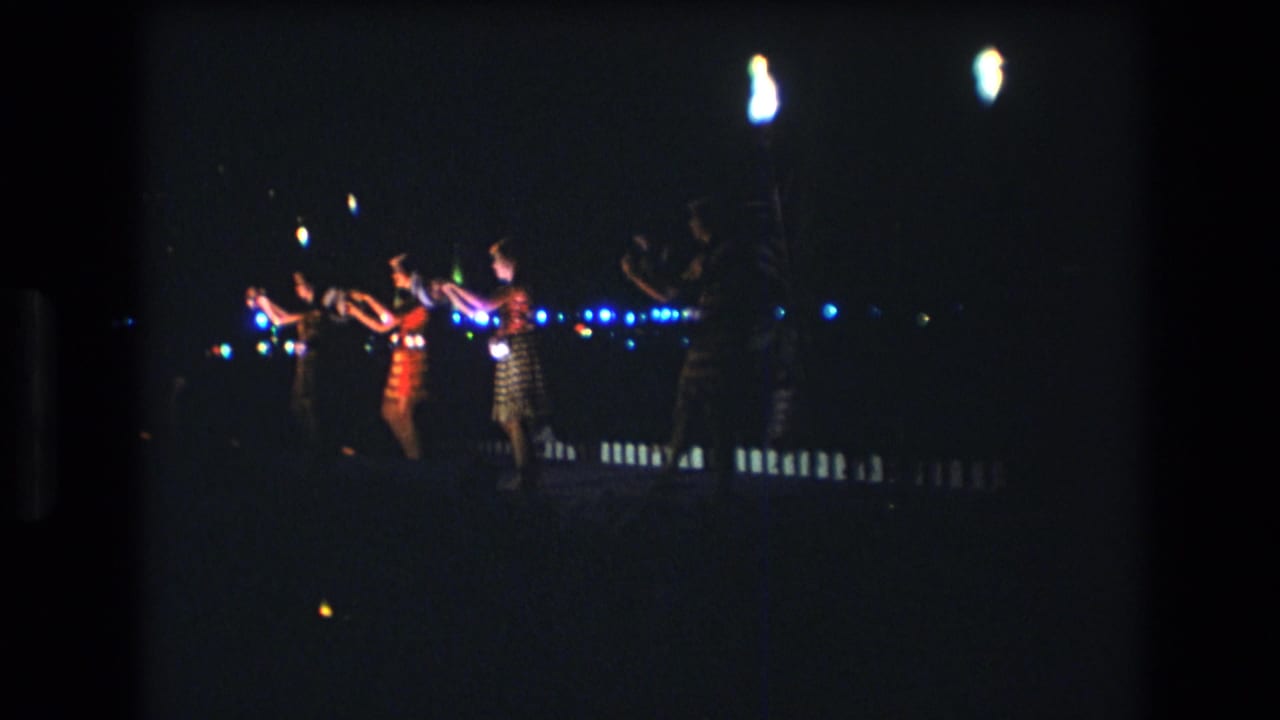 Dancers continue a nighttime performance beside blue lights and bright lamp flares.