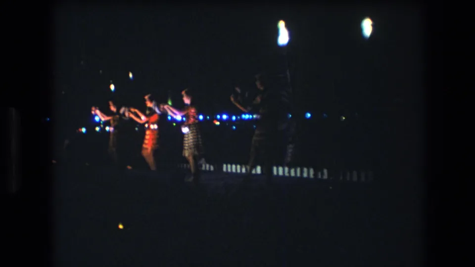 Women in patterned dresses move through a hand-focused dance sequence at night in the 1977 Honolulu 8mm clip, seen from a distant visitor viewpoint.