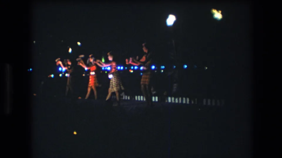 A line of women performs synchronized gestures in a dark outdoor Honolulu setting, with blue lights and bright lamp bloom visible in the 1977 home-movie frame.
