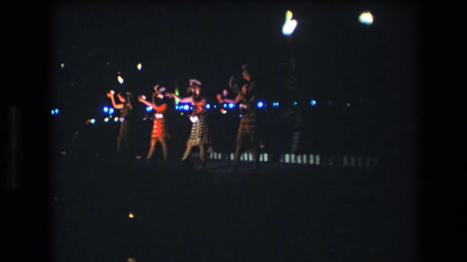 Women continue a synchronized raised-arm dance under bright lamps and blue background lights in a 1977 Honolulu, Oahu 8mm home-movie frame.