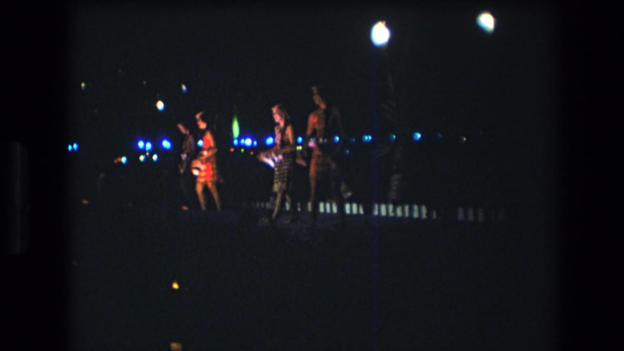 In 1977 Honolulu home-movie footage, women performers in patterned dresses appear on a dark nighttime stage beneath bright lamps with blue background lights.