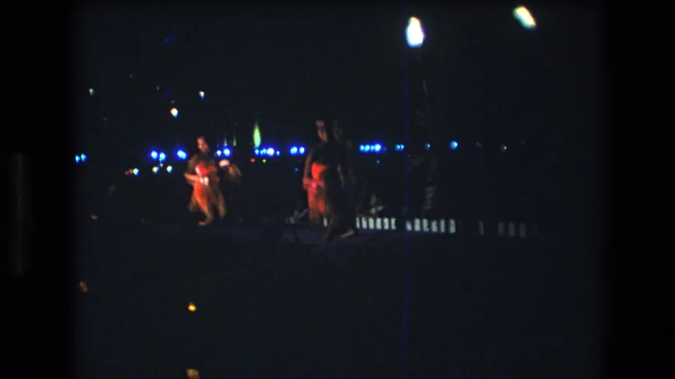 Two women performers move through a low-lit Honolulu stage scene, with one figure holding a bright or reflective prop and blue lights across the background.