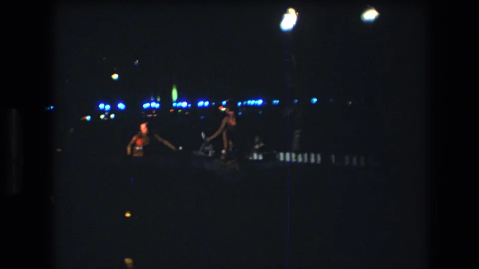 A low-light 8mm frame shows performers near the bottom of the stage area, with illuminated props or reflective objects and distant blue lights behind them.