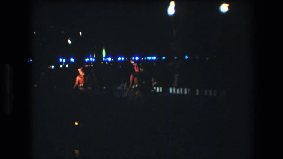 Two women performers are visible at opposite sides of a dim Honolulu stage, framed by overhead lamp glare, blue background lights, and the dark edges of the 8mm scan.