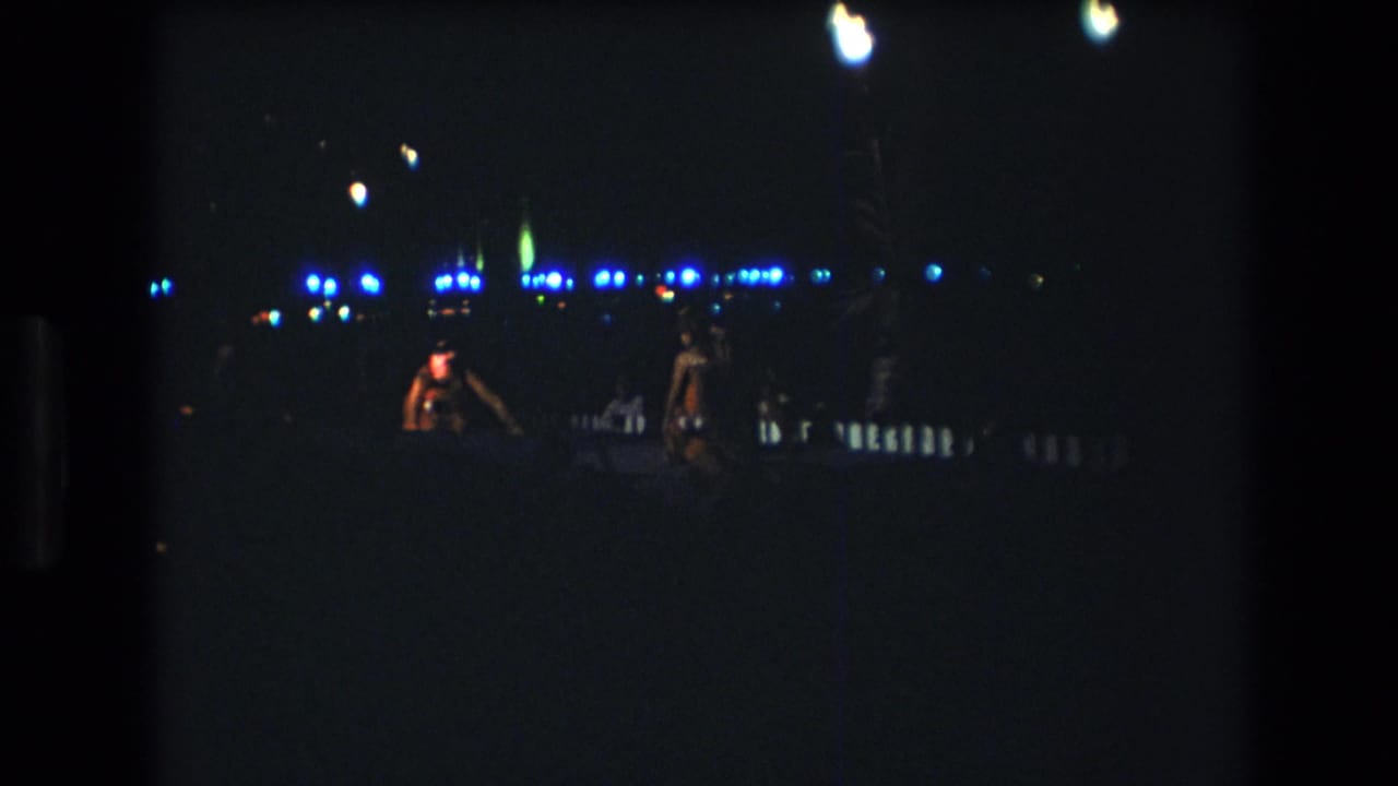 Two adult dancers appear in low light during a 1977 Honolulu, Oahu nighttime party scene, with blue-white lights and small fire glows in the background.