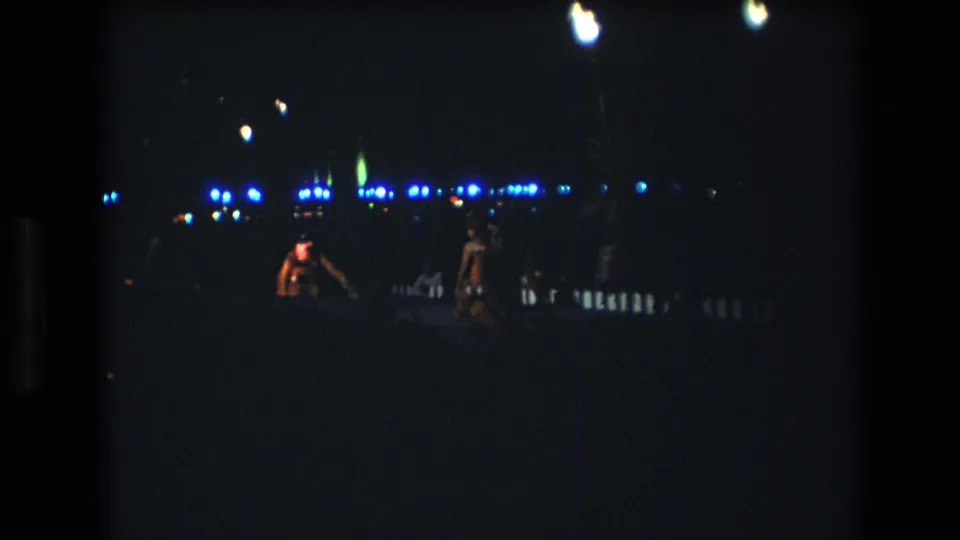 Two adult dancers appear in low light during a 1977 Honolulu, Oahu nighttime party scene, with blue-white lights and small fire glows in the background.