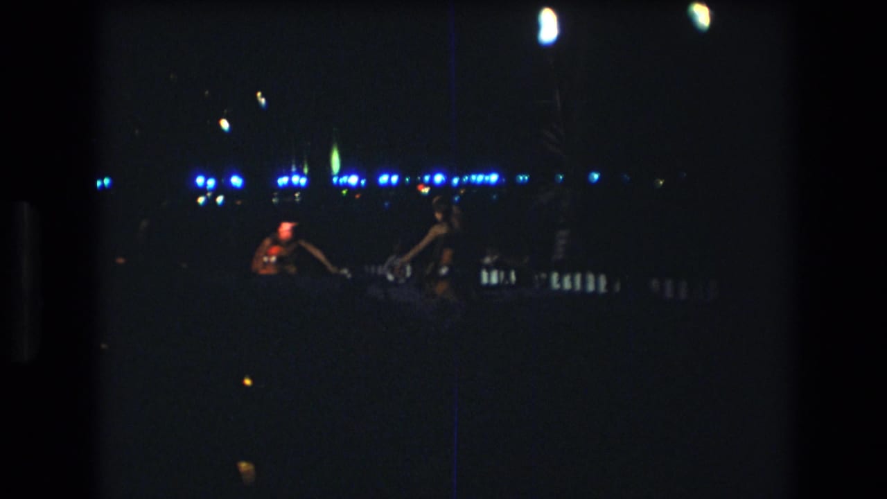 Two dancers move across a very dark party area with lights behind them.