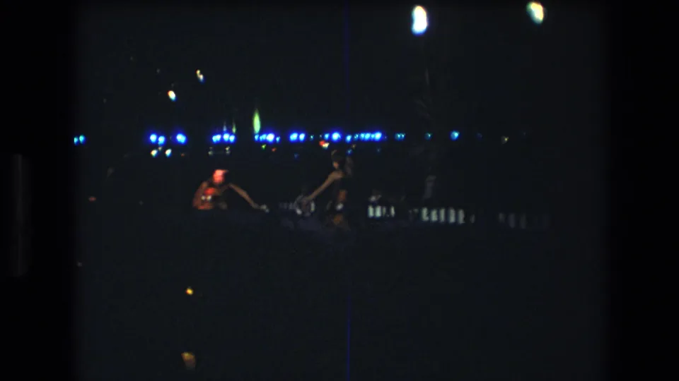 In this 1977 Honolulu, Oahu frame, two adult figures dance in silhouette against a row of blue lights and faint warm firelight.