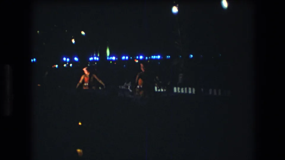 Adult dancers in red-toned tropical attire are visible in a grainy 1977 Honolulu, Oahu nighttime party frame with blue lights across the background.