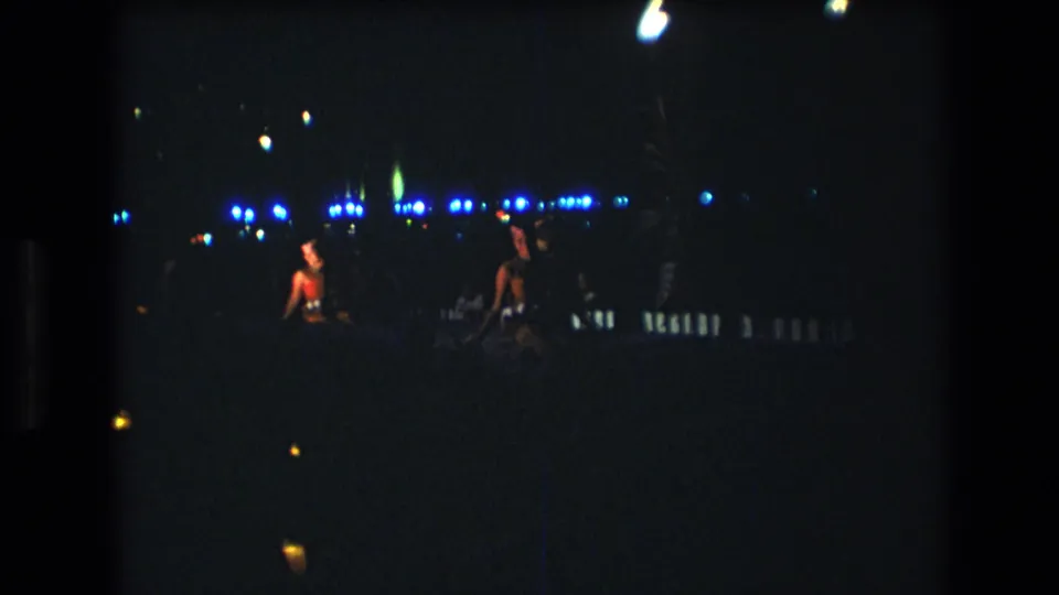 Two adults appear to dance facing one another during the 1977 Honolulu, Oahu nighttime party, with blue light strings and bright overhead bulbs visible through heavy film grain.