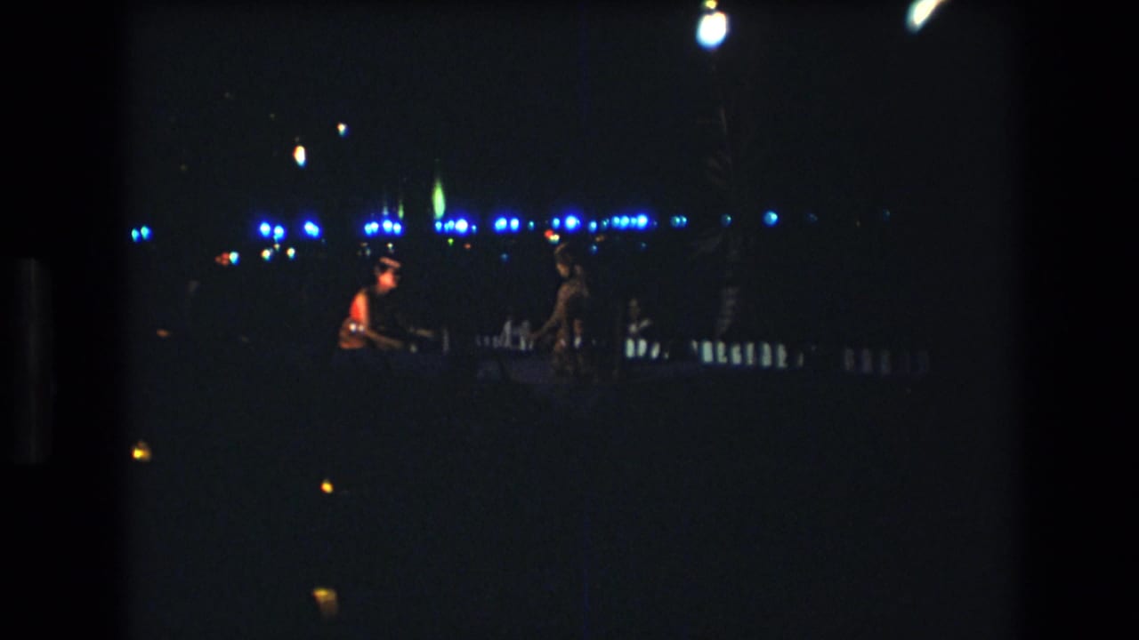 1977 Honolulu home-movie frame showing distant nighttime performers near blue lights, with most details silhouetted in the dark.