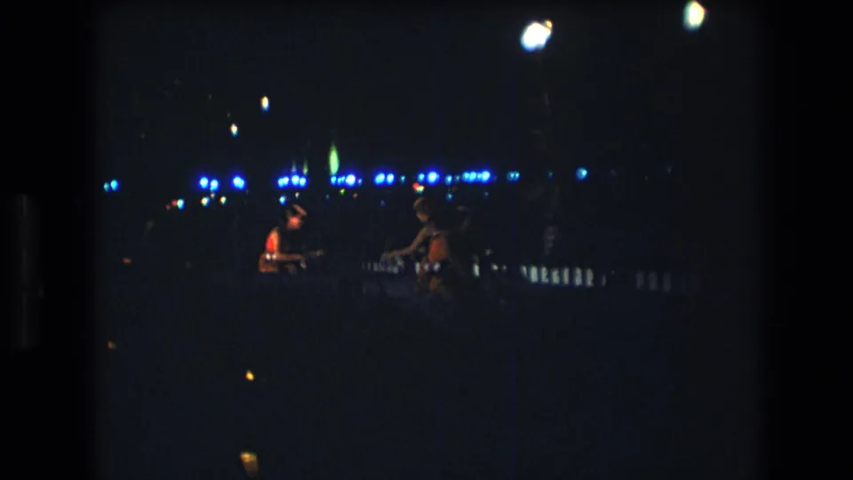 1977 Honolulu 8mm frame with two distant performers or musicians silhouetted beside bright blue event lights in a dark outdoor setting.