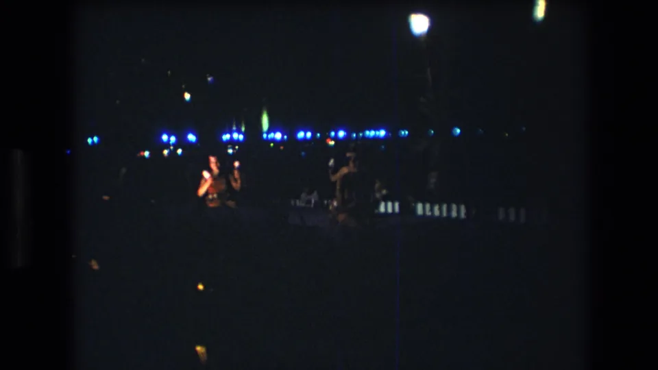 1977 Honolulu home-movie frame showing distant figures during a nighttime performance, with blue lights and warm lamp points across the background.