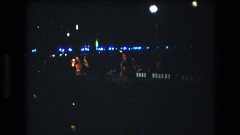 1977 Honolulu 8mm frame showing performers at a dark nighttime music event, with a bright overhead lamp and blue lights behind them.