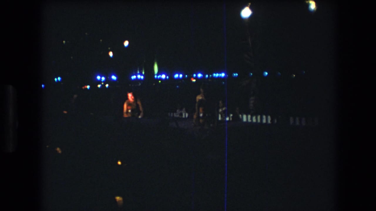 In this 1977 Honolulu 8mm frame, two women appear on a dim nighttime stage with small items in their hands and blue lights behind them.