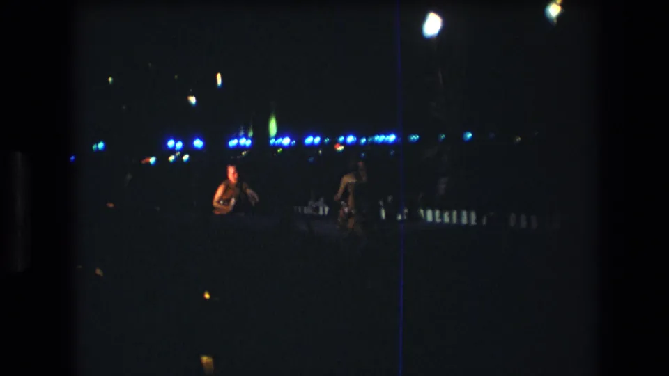 Two women are visible at a distance on a dark Honolulu stage, with blue background lights and bright overhead flares in the 8mm frame.