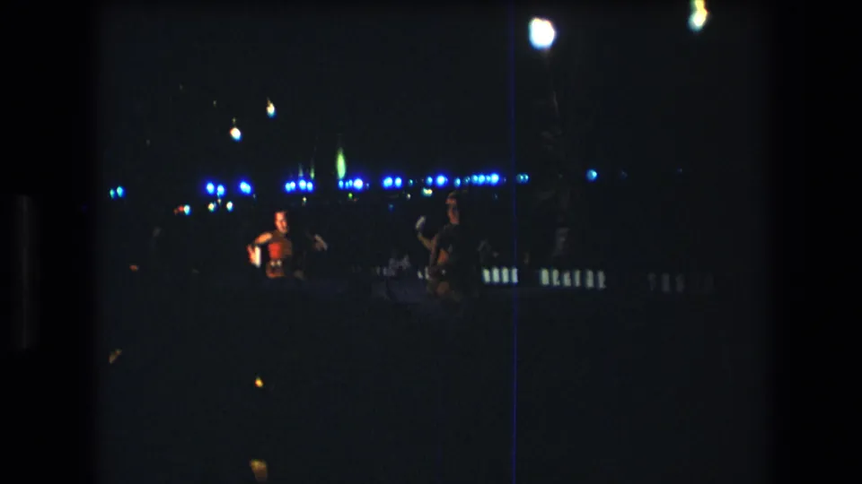A 1977 Honolulu home-movie frame shows a woman holding a small item on a dim stage while another performer stands nearby under blue lights.
