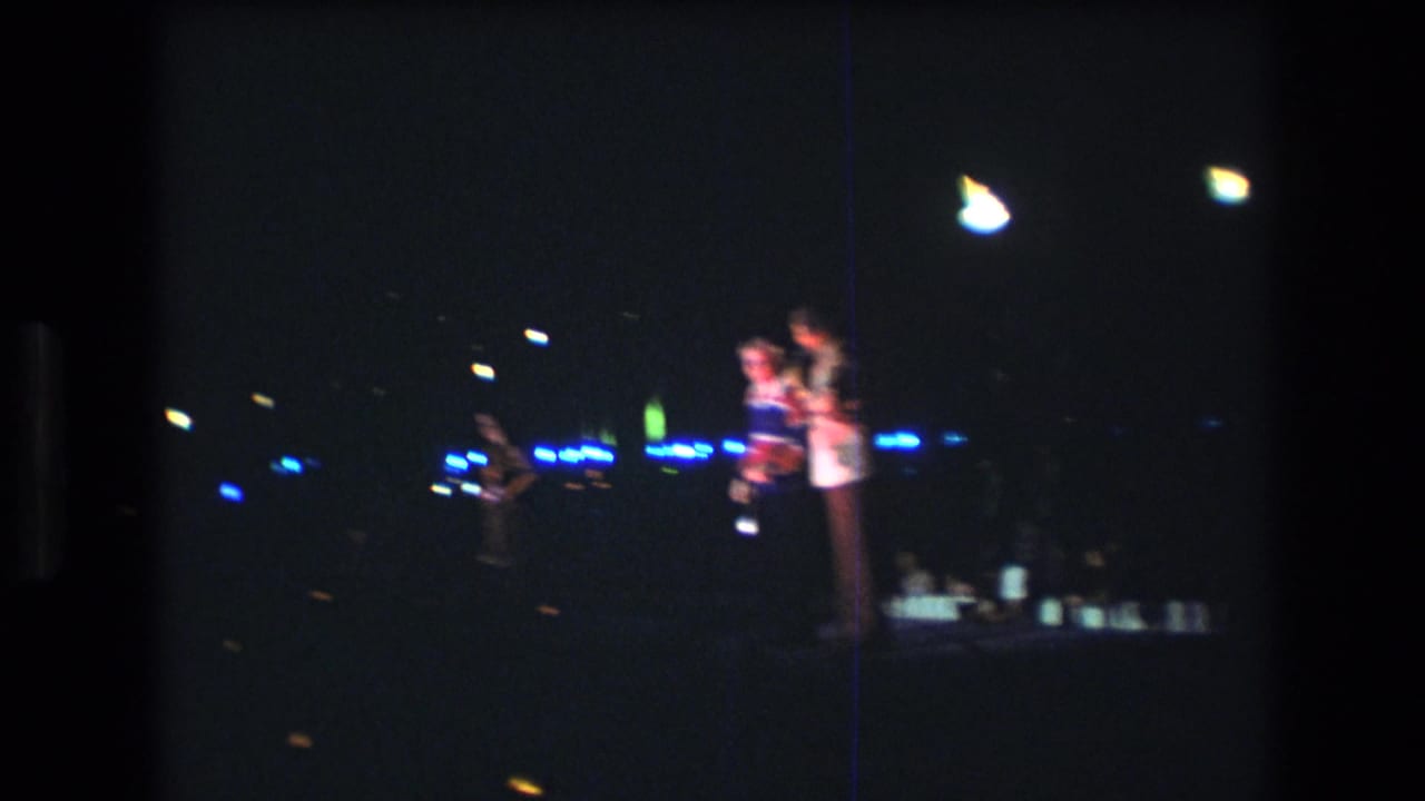 1977 Honolulu 8mm frame showing two people on the right side of a dim stage, one near a microphone, with blue light points and another figure farther back.