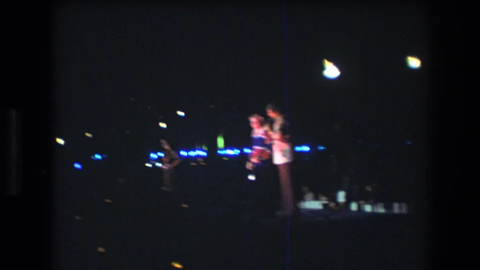 1977 Honolulu 8mm frame showing two people on the right side of a dim stage, one near a microphone, with blue light points and another figure farther back.