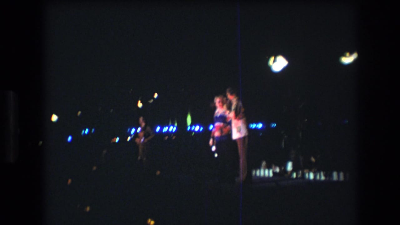 Two people remain at the right of a dark stage while blue lights stretch across the background.