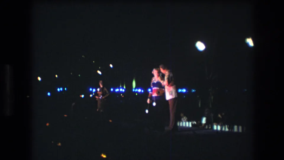 1977 Honolulu 8mm frame showing two people close together on a dimly lit stage, with a microphone, blue background lights, and low-light film softness.
