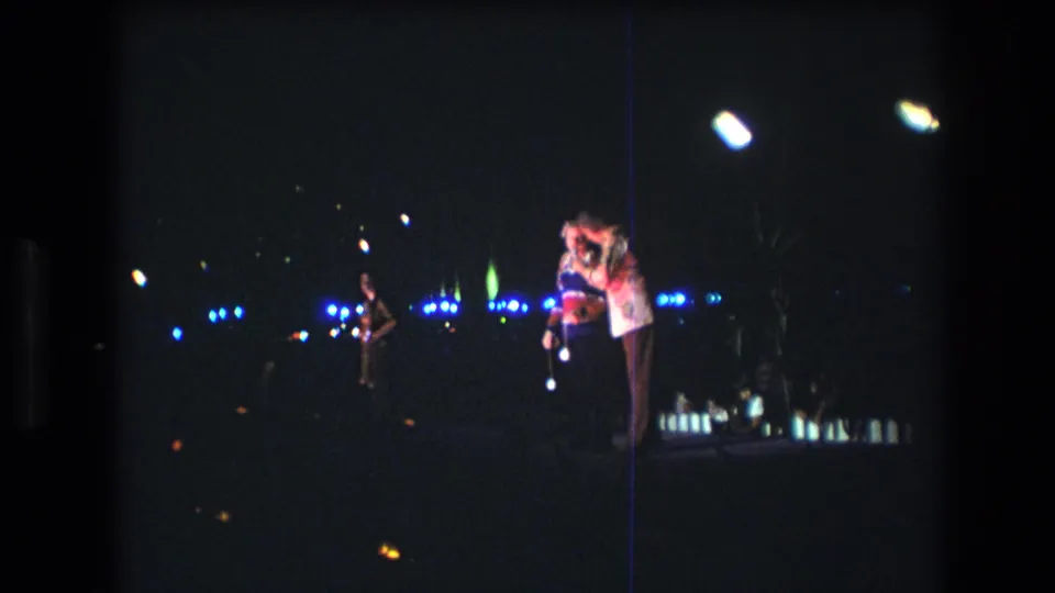 1977 Honolulu 8mm frame with two visible stage figures at right, bright overhead light, a background person, and rows of blue lights in the surrounding darkness.